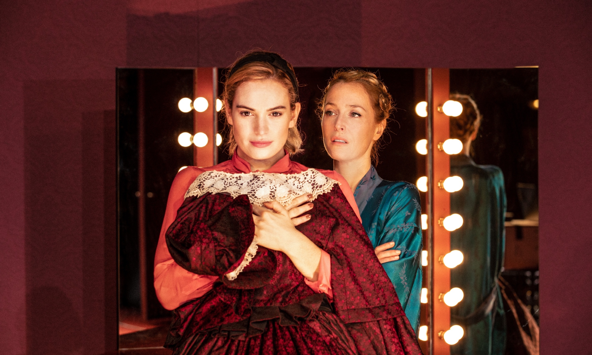 Lily James stands in front of three dressing room mirrors, holding a red dress close to her chest. Behind her, in a blue robe, looking at her, is Gillian Anderson.