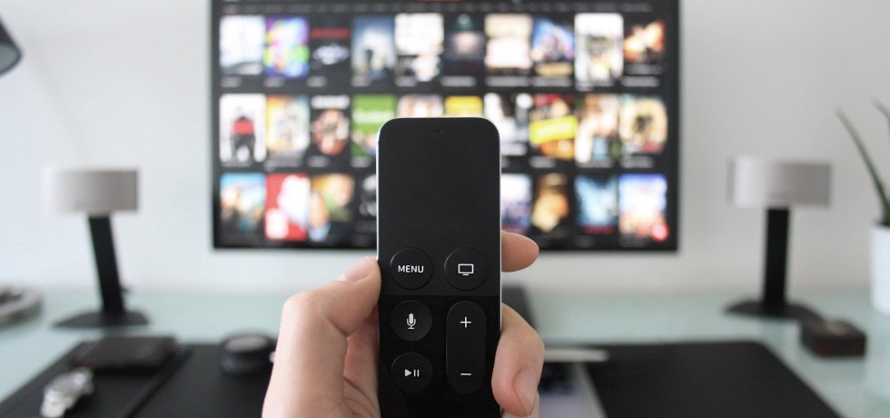 Person holding a TV remote in their hand. Behind that is a TV with the Netflix home screen on it.