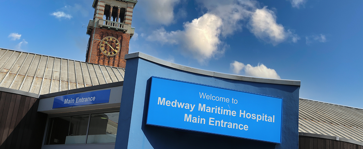 'Welcome to Medway Maritime Hospital Main Entrance' sign on building against blue sky.