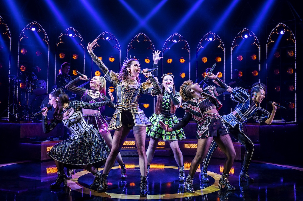 Six women, in glittery Tudor-style costumes, are huddled together and singing on-stage, with their right hands stretched outwards as they do so.