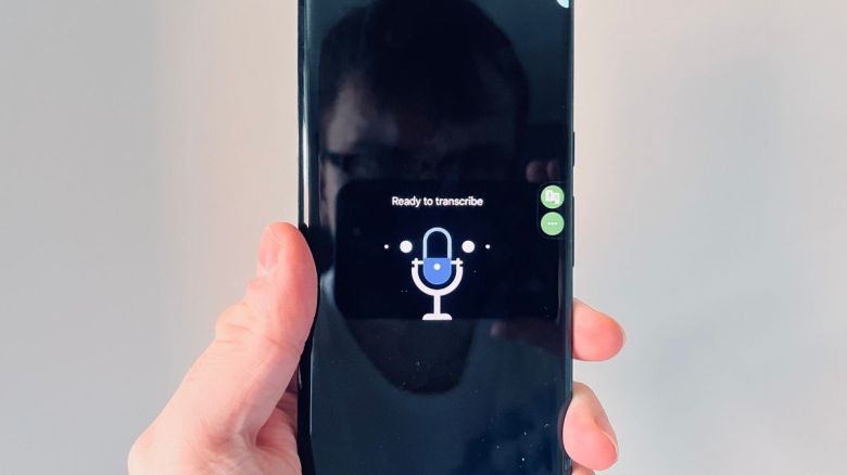 A white hand holds up a Google Pixel phone, showing a blue microphone icon and text which reads: 'Ready to transcribe'.