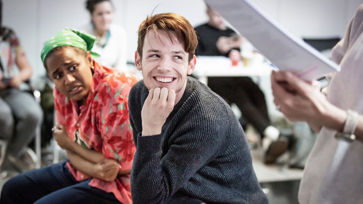 JJ Green, a young white man with short brown hair and a grey jumper, grins at someone off camera. He is sitting down and resting his chin in his left hand. He is in a rehearsal room, and someone holding a script can be seen in the right of the photo. Sitting behind him with a shocked expression on her face is a Black woman in a red floral dress and green bandana, her arms crossed.