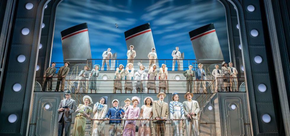 Rows of people sing behind barriers on a large ocean liner set on a theatre stage.