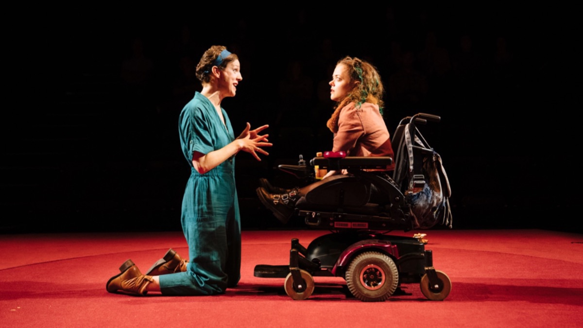 On a red stage is a white woman in a blue playsuit who is kneeling and talking to a short white woman with a brown shirt in an electric wheelchair.
