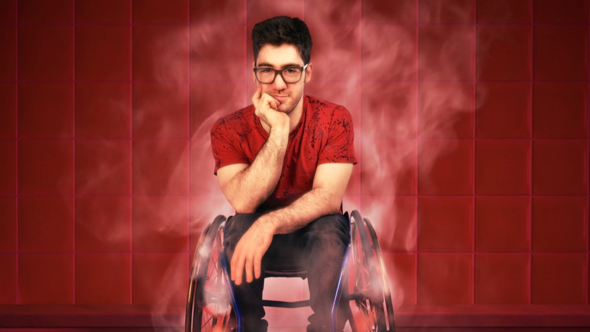 A white man with glasses in a red shirt and wheelchair rests his head in his right hand and smiles. A red wall is behind him and steam is rising from the wheels of his wheelchair.