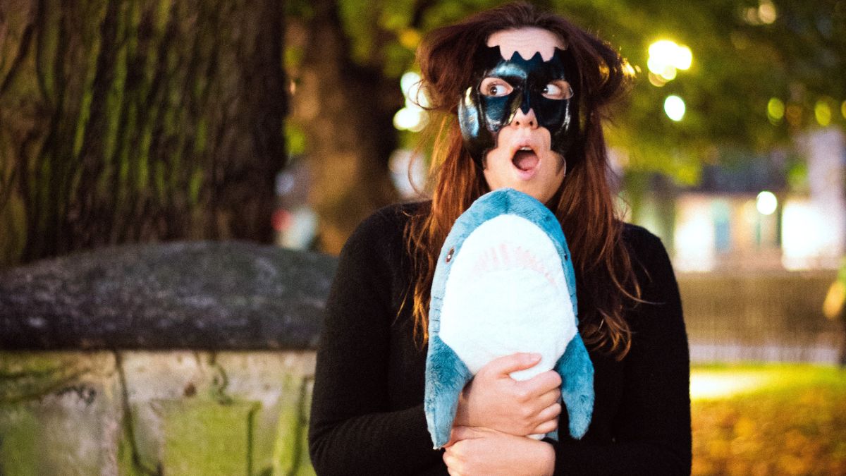 A white woman with long brown hair and a Batman eye mask has a shocked expression on her face as she holds a shark plushie on a park bench.