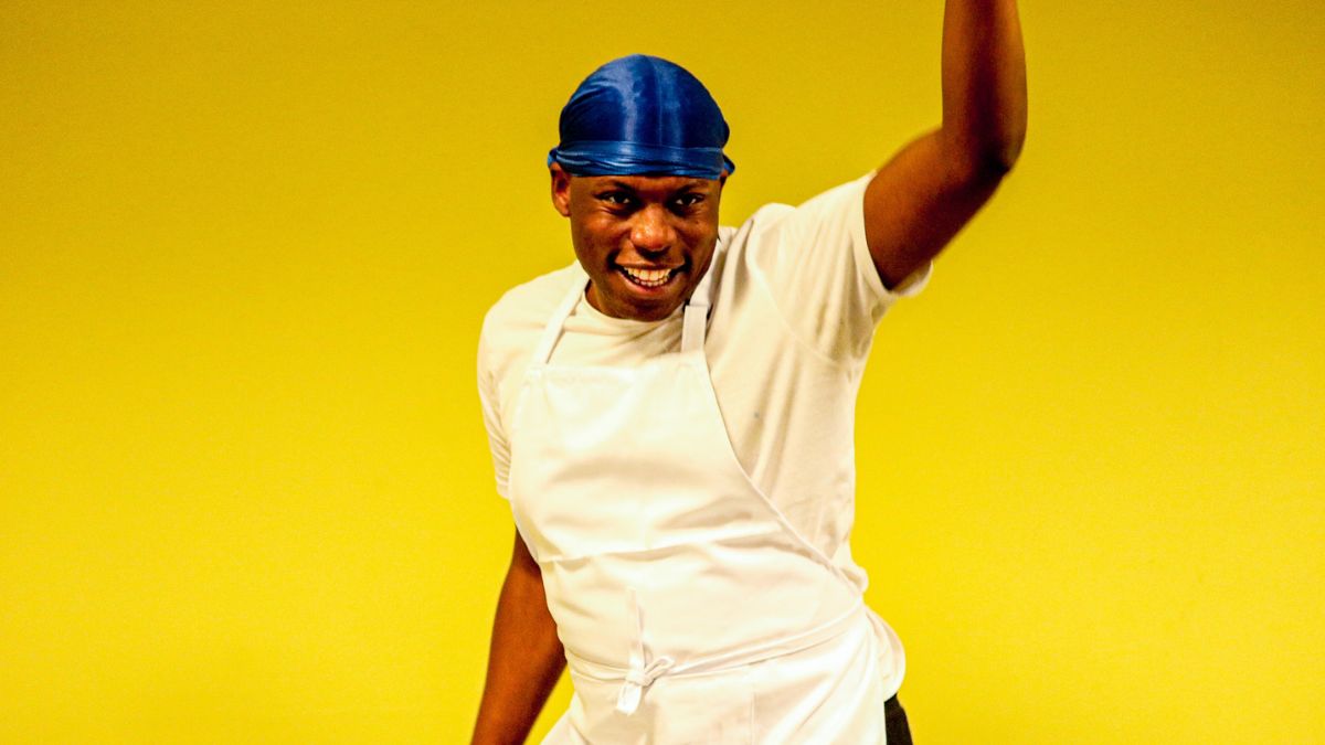A Black man in a white shirt and apron, with a blue durag, smiles and holds one hand up. The background behind them is bright yellow.