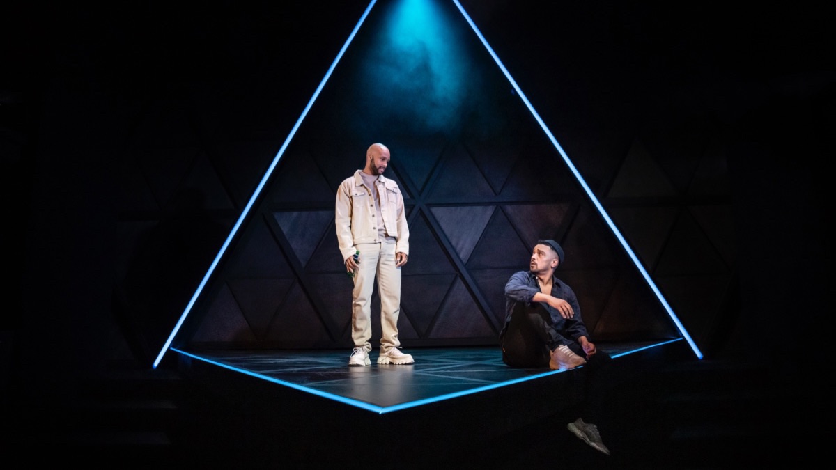 A triangular theatre stage lit up in blue lighting. One Black man in a blue shirt with short black hair. He’s looking up at a Black man in all-white clothing, standing to the left of the stage.