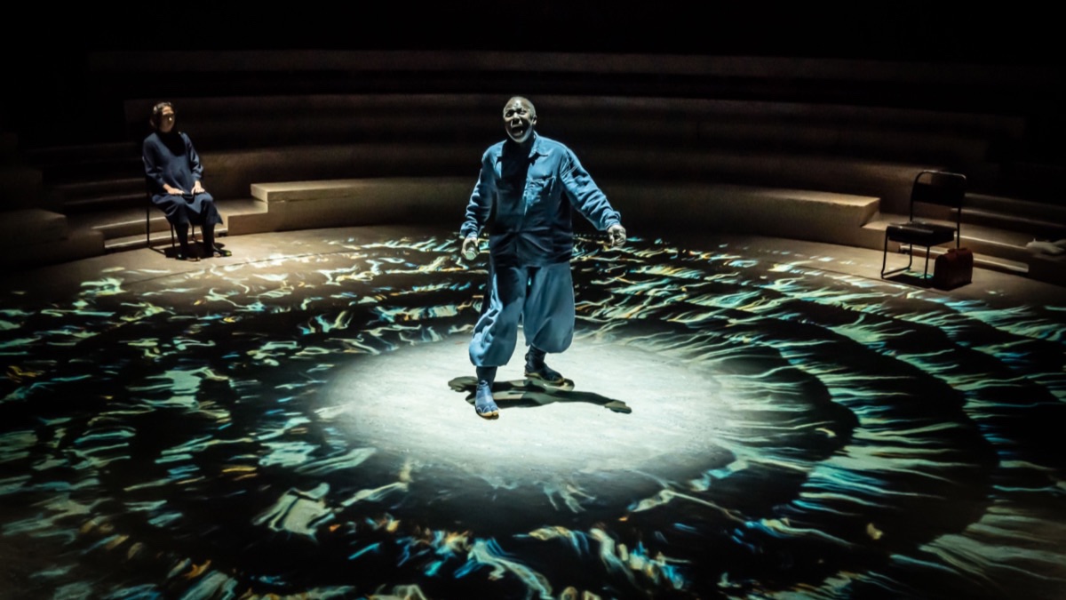A Black man in the centre of a circular stage. He’s in a blue boilersuit, appears to be shouting, and beneath him on the floor of the stage are pulsing blue concentric circles of light.
