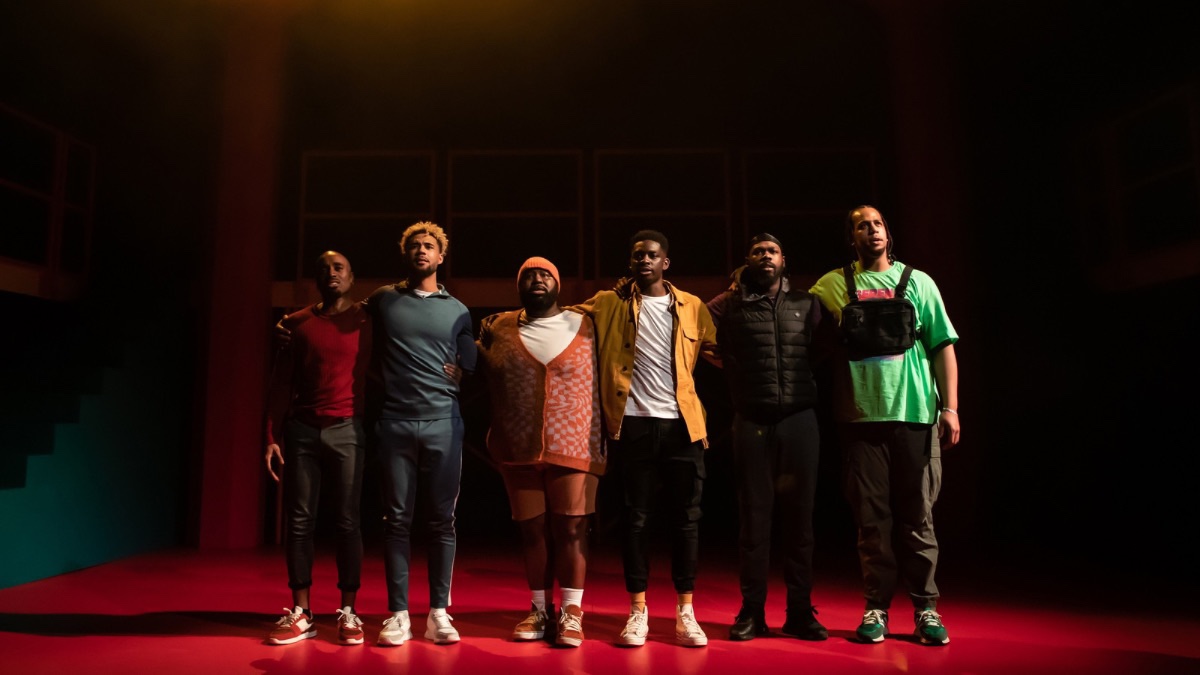Production image from For Black Boys. Six Black men stand in a line with their arms around one another, looking into the audience. They each wear a colour of the rainbow.