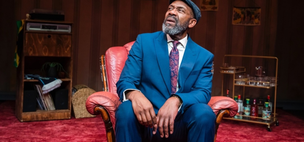 Lenny Henry, a Black man with a short grey beard, is seated on a red armchair on a stage, looking to his right with a look of confusion and concern. He’s wearing a blue suit and grey flat cap, with the set behind him looking like an eccentric living room with wooden cabinets and a red leather carpet.