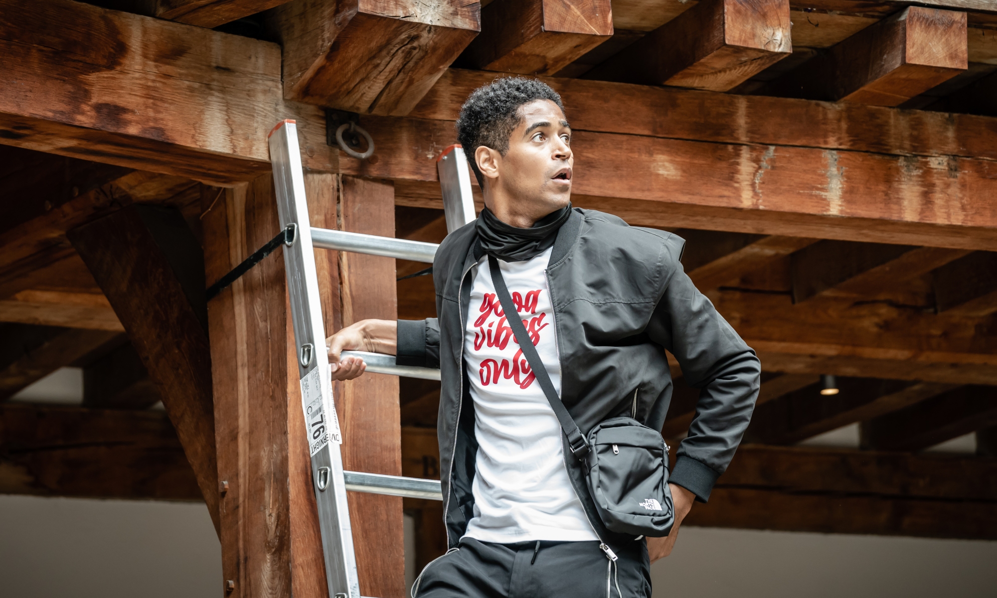 Alfred Enoch, a Black man wearing a black bomber jacket and white shirt with the text 'good vibes only', stands on a ladder affixed to the side of Shakespeare's Globe.