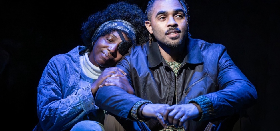 Theatre production image. A Black woman with an afro and eye patch rests her head on the shoulder of a bearded brown man. They’re sat down on the stage.