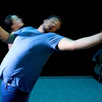 Theatre production photo. In the foreground, a brown man leans backwards slightly, his arms flailing backwards. Behind him, blurred, is a white woman walking to the right.