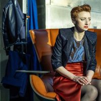 A white woman with a ginger quiff and a black leather jacket sits on an orange armchair, looking concerned and to her left.