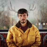 A bearded white man with short brown hair and a yellow jacket sits at the back of a bus, a neutral expression on his face. In the foggy window behind him, out of the vapour, are two antlers coming out of his head.