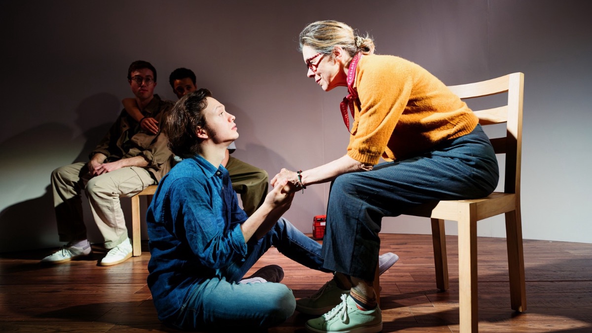 Theatre production photo. A white boy with curly black hair and blue clothing sits on the stage looking up at a white woman with long blonde hair tied in a bun, wearing a yellow jumper and glasses and sitting on a wooden chair.