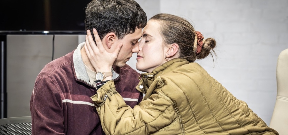 Theatre production image. A white woman in a mustard coat on the right places her left hand on the side of the face of a white man, left, as they go in for a kiss.