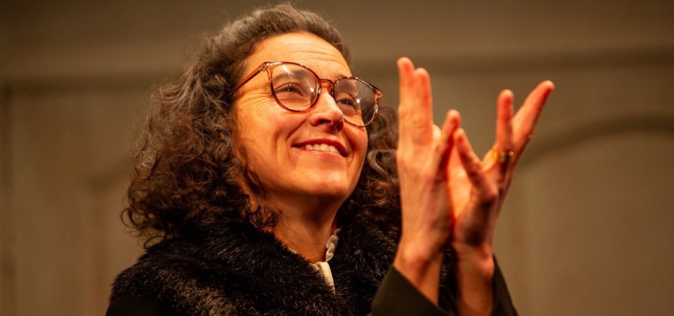 Theatre production image. A white woman with glasses and curly brown hair smiles as she forms a cup with her hands splayed.
