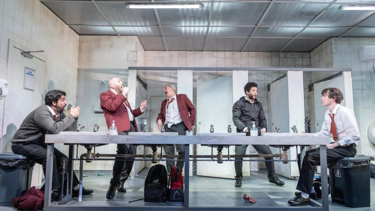 Theatre production photo. Five men playing school boys wear grey and red suits as uniform and are laughing in a public toilet.