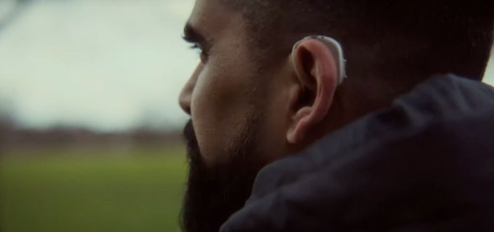 A photo from the side of a bearded brown man looking out at a field, a grey hearing aid in his left ear.